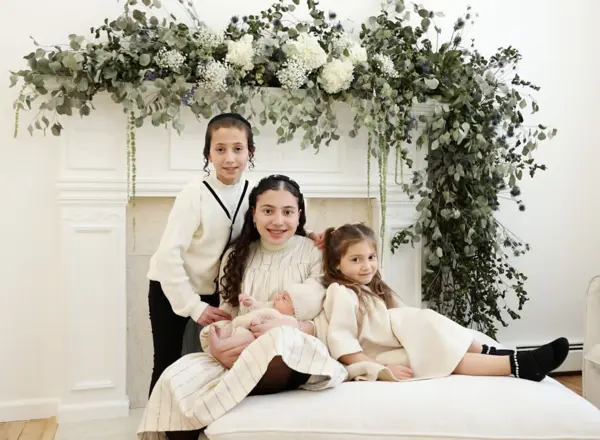 Newborn photography Monsey NY – Big sisters on a white sofa holding their newborn sibling under a floral arch